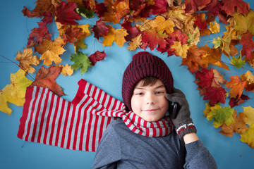 seven years old boy dreaming in autumn. Child in hat and scarf and maple leaves on blue background speaking on the smartphone