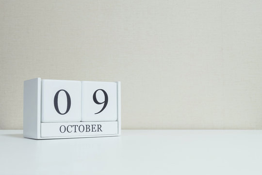 Closeup surface white wooden calendar with black 9 october word on blurred white wood desk and cream color wallpaper in room textured background with copy space