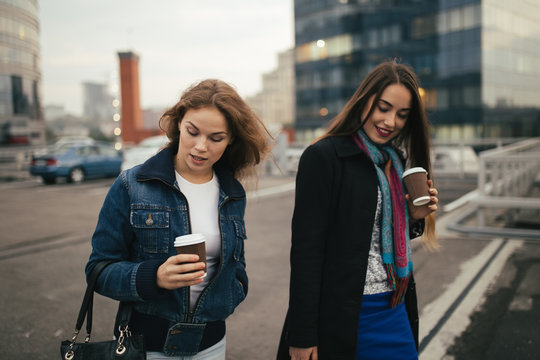 Outdoors fashion portrait of two cheerful girls drinking coffee. Walking in the city.