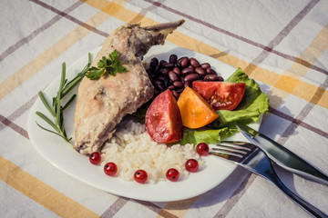 Gourmet roasted rabbit leg with rice and beans, white sauce. Meal is served on white simply plate and checked table cloth, with cutlery. Health and light meal good for diet.