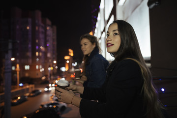 Two young woman friends looking at city street from balcony at night