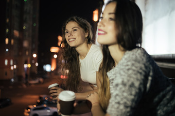 Two young woman friends looking at city street from balcony at night