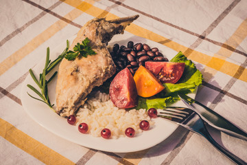Gourmet roasted rabbit leg with rice and beans, white sauce. Meal is served on white simply plate and checked table cloth, with cutlery. Health and light meal good for diet.