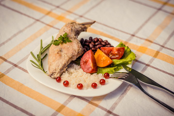 Gourmet roasted rabbit leg with rice and beans, white sauce. Meal is served on white simply plate and checked table cloth, with cutlery. Health and light meal good for diet.