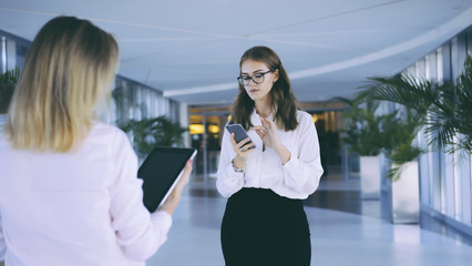Fototapeta premium Two business women standing in the lobby and checking email on the gadgets.Brunette in glasses and a white blouse holding a smartphone,in the foreground a girl with the tablet computer.