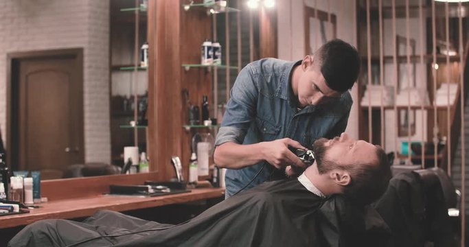 Barber cutting beard with clipper at barbershop 4k video. Styling with electric trimmer razor the client beard in hairdressing salon
