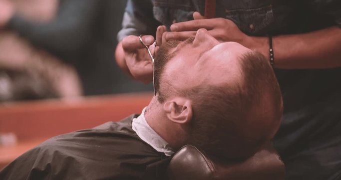 Barber cutting client beard with scissors in barbershop 4k close-up video. Hairdressing of lumberjack bearded man