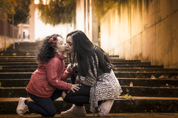 Niña y adolescente, hermanas felices dándose un beso en las escaleras con el sol al fondo y a contraluz.