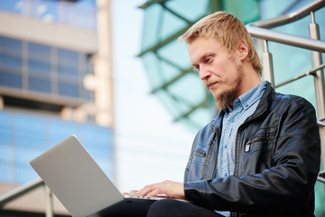The blond guy with a beard in a leather jacket and shirt, sitting on the steps and typing on computer in the background of city buildings