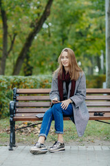 Beautiful stylish young woman in a warm scarf blue jeans trendy shoes and cozy coat sitting alone on bench waiting in fall city park