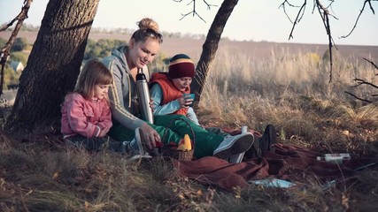 Full Length of Woman with Two Children Sitting Underneath Tree with Warm Beverage in Metal Thermos - Stopping to Enjoy a Hot Drink Outdoors in Early Evening. - Powered by Adobe