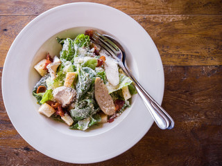 Top view of the delicious Caesar salad that contains green cos vegetable, crispy bacon, crouton and grilled chicken sprinkle over with Parmesan cheese in a white dish that is placed on a wooden table.