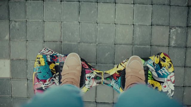Shooting from the first person: Women's shoes on bright and colorful gyroscooter. Woman in jeans goes straight on paved track. A popular means of transportation Hoverboard.