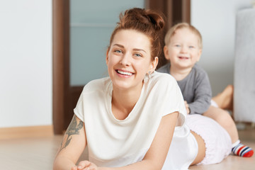 Nice family shot of young mother and her little son playing on the floor at home. Attractive Caucasian woman in white top, lying on her belly. Smiling kid straddling happily on her shinny mommy.