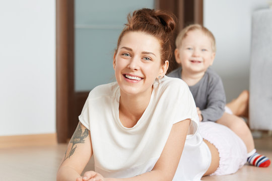 Nice family shot of young mother and her little son playing on the floor at home. Attractive Caucasian woman in white top, lying on her belly. Smiling kid straddling happily on her shinny mommy.