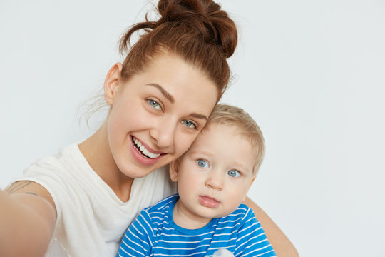 Nice selfie of mother and child on white background. Young Caucasian woman with blue eyes, smiling, holding in her arms little blond boy in blue stripped T-shirt. Happy family self-portrait.