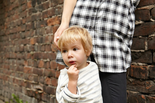 Close-up portrait of little blond kid and his mother near brick wall. Cropped image of pensive male child and figure of loving mom in checked shirts and trousers. Family walk at city back streets.