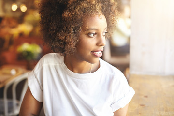 Profile shot of cute young dark-skinned student girl dressed in stylish top spending free time at university canteen, waiting for her groupmates to join her, sitting at window and looking outside