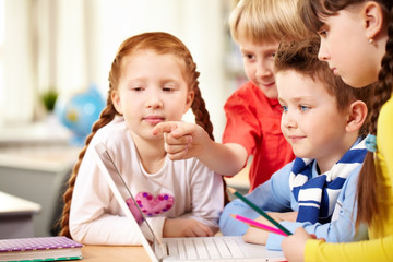 Fototapeta premium Four schoolchildren sitting at table and looking at laptop screen