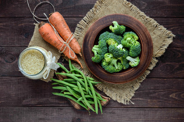 Broccoli (cabbage) pieces in a clay bowl on a wooden background with asparagus, rice and carrot in the background. Set of products for vegan dishes. The top view