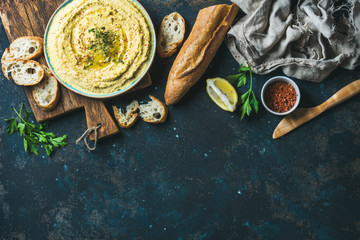 Homemade hummus dip in blue ceramic bowl with fresh baguette, lemon, herbs and spices over dark...