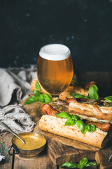 Glass of wheat unfiltered beer and homemade grilled sausage dogs in baguette with mustard, caramelised onion and herbs on serving board over rustic wooden background, selective focus, copy space