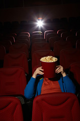 Fototapeta premium Young man hiding behind popcorn bucket while watching horror film
