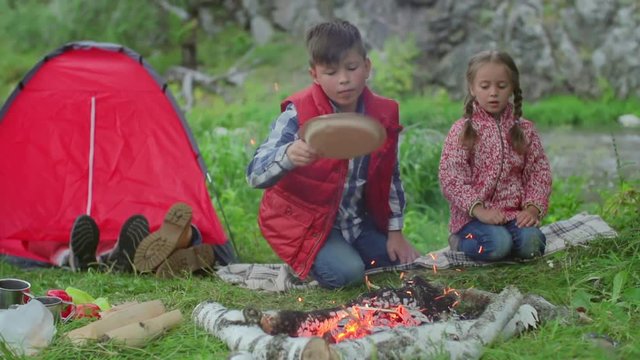 Boy blowing air with paper plate on campfire as his little sister looking, feet of parents sleeping in tent in background  