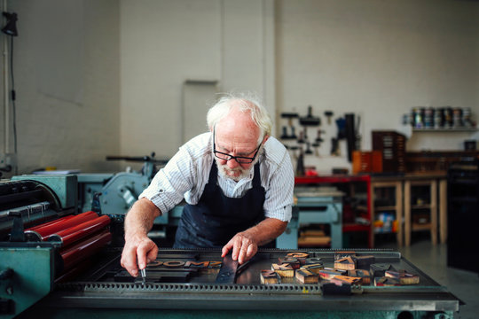 Senior craftsman bending over and using wooden letters and letterpress machine in book arts workshop