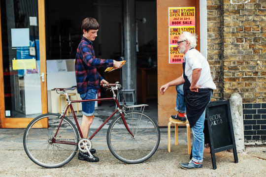 Young man with pushbike looking at post and standing with senior craftsman outside print workshop