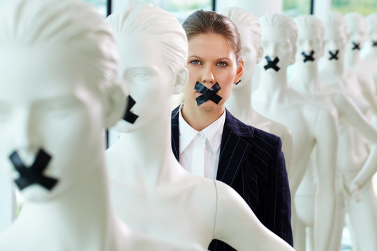 A Woman Standing In Line Of Mannequins With Taped Mouth