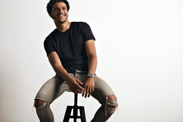 Happy smiling black young man sitting comfortably on a black stool wearing gray jeans and an unlabeled black t-shirt isolated on white