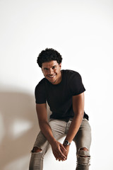 Smiling athletic young African American man in blank black cotton t-shirt and jeans sitting at a white wall