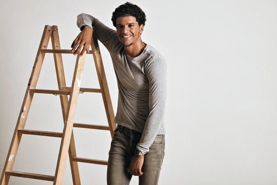 Smiling relaxed tall muscular young model wearing a plain heather gray longsleeve t-shirt and slim gray jeans leaning on a wooden stepladder isolated on white.