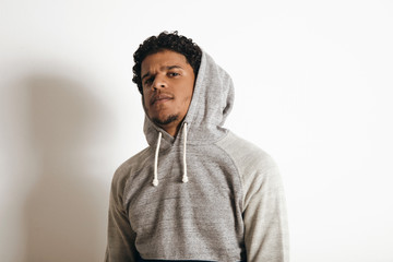 Close up of latino handsome guy wearing hood of his blank grey sweater looking weirdly on camera, isolated on white