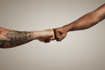 Two men one light skinned with tattoos and another dark skinned do a fist bump against white wall close up