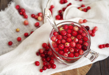 Fresh berries of a forest red cranberry in transparent glass with water among the scattered berries on a white linen cloth on a wooden surface of a table