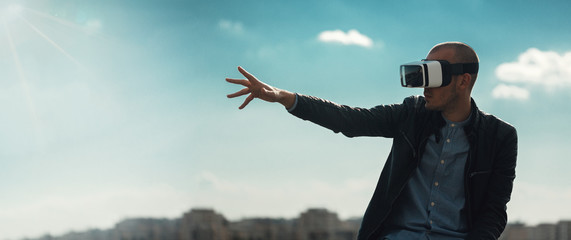 Excited young man using a pair of VR glasses above the city with the blue sky background amazed by...