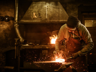 The blacksmith forging the molten metal on the anvil in smithy - obrazy, fototapety, plakaty