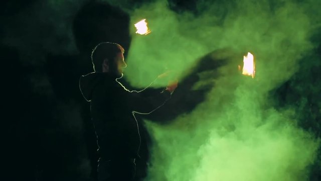 Side view of man performing 2-beat weave trick with fire poi with green smoke in the background at night