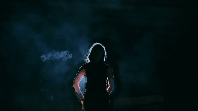 Woman standing in cloud of smoke in the darkness and rotating burning torch behind her back in slow motion