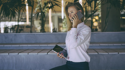 Fototapeta premium View from the side, a woman with blonde hair in a light pink shirt sitting and talking on a cell phone, in her hand she holds a tablet computer with a blank screen.Film effect, blurred background.