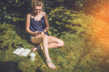 Girl with blond hair sitting under tree on green lawn and using gadget.Next on the grass are notebook,pen and cup of coffee.Young woman in top and shorts use a smartphone while sitting on the grass.
