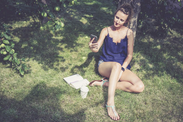 Young woman sitting in garden under apple tree and uses digital gadget.Girl,dressed in blue top and short denim shorts,sitting on grass and surf Internet on smartphone.Near notebook and cup of coffee.