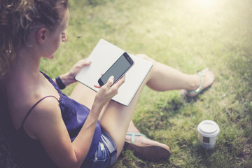 View from behind,girl holding smartphone with blank screen,while sitting on lawn under tree.In her hand notebook,near cup coffee.Woman sitting on grass and check email on smartphone.Girl using gadget.