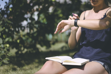 Summer sunny day.Close-up of a girl using smartwatch. A young woman wearing a blue top and denim shorts sitting on the lawn under a tree, and clicks on the screen digital gadget, notebook is on knees.