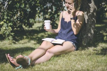 Sunny summer day,girl dressed in blue silk top and denim shorts sitting on lawn under tree talking on cell phone and drinking coffee while reading the entries in the notebook.On the wrist smartwatch.