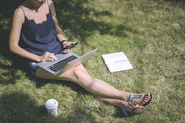 Sunny summer day,young woman uses laptop while sitting on the lawn under tree.In one hand she holds smartphone,on the grass near the girl is notebook and cup of coffee.Girl using gadget.