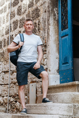 Stylish tourist. Man dressed in a white shirt and blue shorts with a blue backpack over his shoulder. Standing on the steps of an old European city.