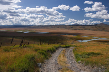 Road along the border line on a highland mountain plateau with the green grass at the background of the valley of white river under a blue sky with white clouds, Plateau Ukok, Altai, Siberia, Russia